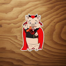 Load image into Gallery viewer, Rudy’s Bar &amp; Grill Devil Pig sticker in a Dracula costume on a plain rustic wood background.