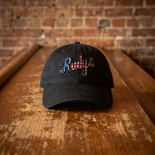 Load image into Gallery viewer, A black baseball cap with 'Rudys' embroidered in red, white, and blue script on the front, resting on a rustic wooden table with a brick wall background.