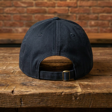 Load image into Gallery viewer, A dark navy blue baseball cap shown from the back, resting on a rustic wooden table with a brick wall background.
