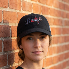 Load image into Gallery viewer, A woman wearing a black baseball cap with 'Rudys' embroidered in cursive script across the front, posed against a warm-toned brick wall.