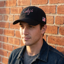 Load image into Gallery viewer, A man wearing a black baseball cap with 'Rudys' embroidered in pink and white script on the front, standing against a red brick wall. The cap also features a small American flag patch on the side.