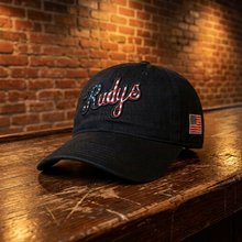 Load image into Gallery viewer, A black baseball cap with 'Rudys' embroidered in red and white script on the front, resting on a rustic wooden table against a warm brick wall background.
