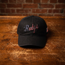 Load image into Gallery viewer, A black baseball cap with 'Rudys' embroidered in cursive across the front, resting on a rustic wooden table with a brick wall background.