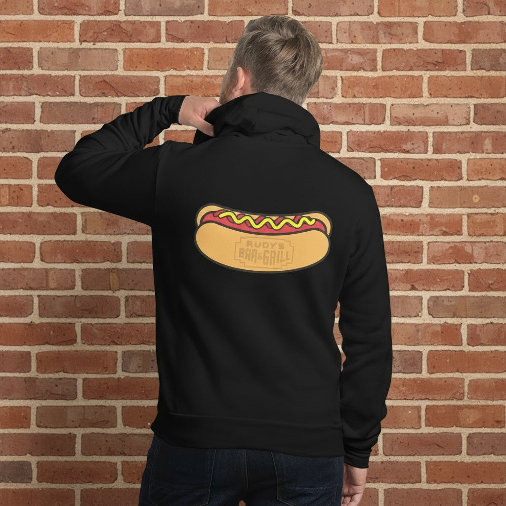 Person wearing Navy Black hoodie featuring a large Rudy’s Bar & Grill hot dog logo on the back, displayed against a reddish-brown textured brick wall background. The hoodie is centered and well-lit, creating a realistic product mockup for Rudy’s Bar & Grill merchandise