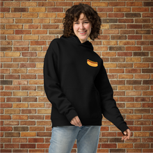 Load image into Gallery viewer, Front view of Rudy’s Bar &amp; Grill Hot Dog Neon Hoodie in black, worn by a smiling model, featuring the hot dog logo on the chest, displayed against a brick wall background.