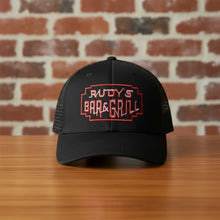 Load image into Gallery viewer, Front view of Rudy’s Bar &amp; Grill black mesh trucker hat with red embroidered logo, displayed on a wooden bar in front of a brick wall