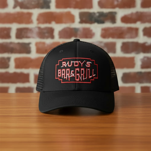 Front view of Rudy’s Bar & Grill black mesh trucker hat with red embroidered logo, displayed on a wooden bar in front of a brick wall