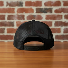 Load image into Gallery viewer, Rear view of Rudy’s Bar &amp; Grill black mesh trucker hat, showing the adjustable snapback closure and breathable mesh back, displayed on a wood bar with a brick wall background