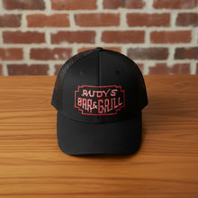 Load image into Gallery viewer, Top-down front view of Rudy’s Bar &amp; Grill black mesh trucker hat with red embroidered logo, placed on a wooden bar in front of a brick wall