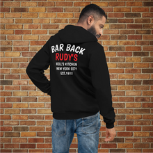 Load image into Gallery viewer, Male model wearing Rudy’s Bar Back Hoodie in black, showing “BAR BACK RUDY’S HELL’S KITCHEN NEW YORK CITY EST. 1933” text on the back, brick wall background