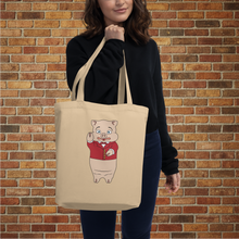 Load image into Gallery viewer, Woman holding Rudy’s Bar &amp; Grill Classic Pig Eco Tote Bag featuring Baron Von Swine mascot, brick wall background