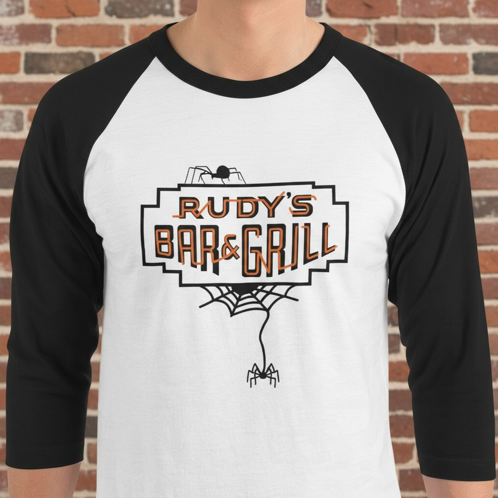 Close-up of a man wearing a white and black raglan 3/4 sleeve shirt with Rudy’s Bar & Grill Halloween spider web logo, standing in front of a reddish-brown brick wall.
