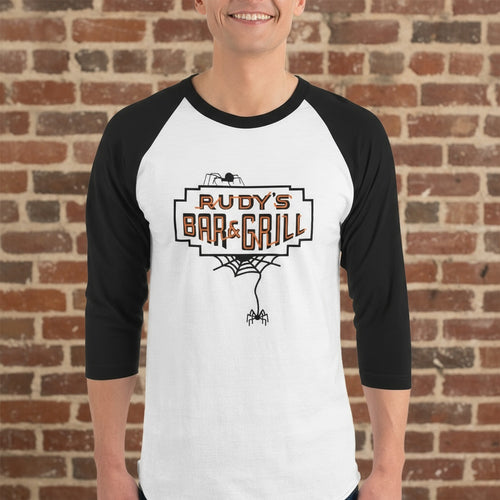 Man wearing a white and black raglan 3/4 sleeve shirt with Rudy’s Bar & Grill Halloween spider web logo, standing in front of a reddish-brown brick wall.
