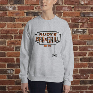 Woman wearing a sport grey crew neck sweatshirt with Rudy’s Bar & Grill Halloween logo and hanging spider, standing in front of a reddish-brown brick wall.