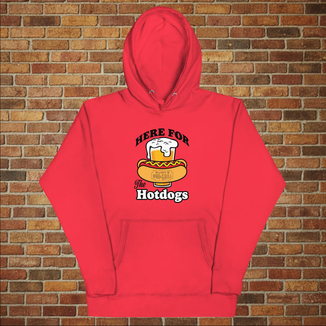 Rudy’s Bar & Grill “Here For the Hotdogs” hoodie in red, front view, featuring beer and hot dog graphic with Rudy’s logo, flat lay on brick wall background.
