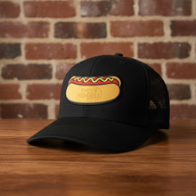 Load image into Gallery viewer, Black mesh-back trucker hat with Rudy’s Bar &amp; Grill hot dog logo patch, displayed on a wooden surface with a reddish-brown brick wall background. Product mockup for Rudy’s Bar &amp; Grill