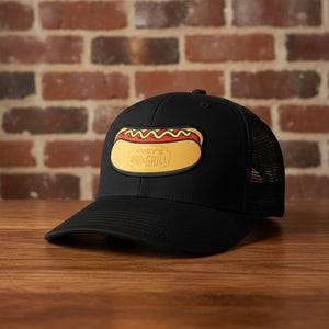 Black mesh-back trucker hat with Rudy’s Bar & Grill hot dog logo patch, displayed on a wooden surface with a reddish-brown brick wall background. Product mockup for Rudy’s Bar & Grill