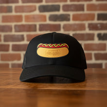 Load image into Gallery viewer, Front view of a black mesh trucker hat with a Rudy’s Bar &amp; Grill hot dog patch logo, displayed on a wooden bar with a brick wall background.