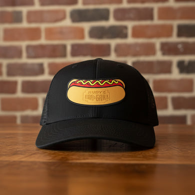 Front view of a black mesh trucker hat with a Rudy’s Bar & Grill hot dog patch logo, displayed on a wooden bar with a brick wall background.