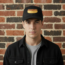 Load image into Gallery viewer, Man wearing a black mesh trucker hat with a Rudy’s Bar &amp; Grill hot dog patch on the front, standing against a rustic brick wall.