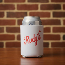 Load image into Gallery viewer, Can koozie with Rudy’s Bar &amp; Grill red script logo on a white sleeve, placed on a wooden table with a brick wall background