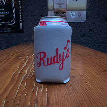 Load image into Gallery viewer, Rudy’s Bar &amp; Grill white can koozie with red script logo, displayed on a rustic wooden bar surface with a dark wall background