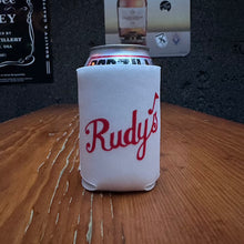 Load image into Gallery viewer, Rudy’s Bar &amp; Grill white can koozie with red script logo, shown on a rustic wooden bar with a dark textured wall and beverage posters in the background