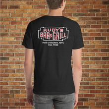 Load image into Gallery viewer, Back view of a black heather t-shirt featuring the Rudy’s Bar &amp; Grill neon sign logo and “Hell’s Kitchen, NYC Est. 1933,” modeled in front of a brick wall.
