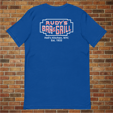 Load image into Gallery viewer, Back view of a true royal blue t-shirt featuring the Rudy’s Bar &amp; Grill neon sign logo with “Hell’s Kitchen, NYC Est. 1933,” modeled in front of a brick wall.