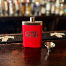 Load image into Gallery viewer, Rudy’s Bar &amp; Grill red Yeti flask with matching funnel displayed on a bar counter, with liquor bottles blurred in the background