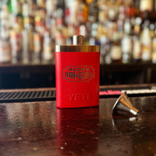 Load image into Gallery viewer, Side view of Rudy’s Bar &amp; Grill red Yeti flask and funnel on a classic bar top, with a colorful bottle display in the background