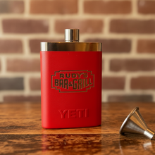 Load image into Gallery viewer, Close-up of Rudy’s Bar &amp; Grill red Yeti flask and metal funnel on a rustic wood table, highlighting the engraved logo and stainless steel cap with a blurred brick wall background