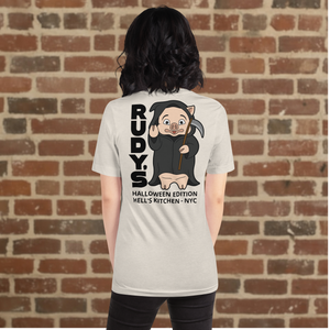Woman wearing Rudy’s Bar & Grill Halloween Edition heather dust t-shirt showing Baron Von Swine dressed as the Grim Reaper with vertical "RUDY’S" text and "Halloween Edition Hell’s Kitchen - NYC" on the back, standing against a brick wall background