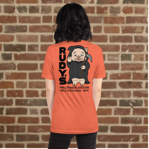 Woman wearing Rudy’s Bar & Grill Halloween Edition heather orange t-shirt showing Baron Von Swine dressed as the Grim Reaper with vertical "RUDY’S" text and "Halloween Edition Hell’s Kitchen - NYC" on the back, standing against a brick wall background