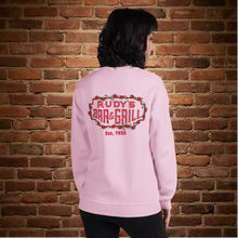 Load image into Gallery viewer, Person showing the back of a light pink crew neck sweatshirt with Rudy’s Bar &amp; Grill neon sign design and “Est. 1933,” standing in front of a brick wall
