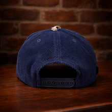 Load image into Gallery viewer, Back view of Rudy’s navy corduroy hat showing adjustable strap, on wood surface with brick wall background