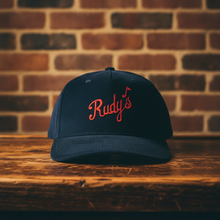 Load image into Gallery viewer, Close-up of Rudy’s navy blue corduroy hat with red script logo, displayed on a rustic wood bar in front of a brick wall