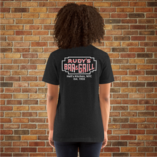Load image into Gallery viewer, Woman modeling the back of a black heather Rudy’s Bar &amp; Grill t-shirt with a neon sign logo and “Hell’s Kitchen, NYC Est. 1933” design, standing in front of a brick wall.