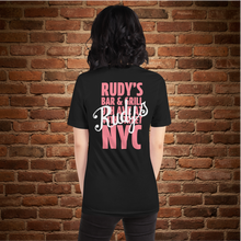 Load image into Gallery viewer, Woman wearing Rudy’s Bar &amp; Grill Valentine’s Day NYC Edition T-shirt, back view – bold pink NYC logo and script on black shirt, brick wall background