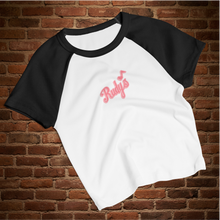 Load image into Gallery viewer, Flat lay of Rudy’s Bar &amp; Grill women’s micro rib raglan baby tee in white with black sleeves, featuring Rudy’s script logo, displayed on a rustic brick wall background