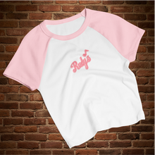 Load image into Gallery viewer, Flat lay of Rudy’s Bar &amp; Grill women’s micro rib raglan baby tee in white with pink sleeves, featuring Rudy’s script logo, displayed on a rustic brick wall background