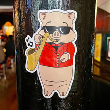 Load image into Gallery viewer, Baron Von Swine jazz pig sticker from Rudy’s Bar &amp; Grill displayed on a bar post, featuring the mascot in sunglasses and a red jacket playing saxophone
