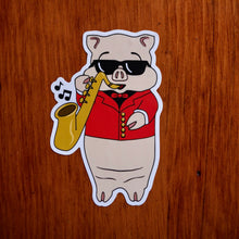 Load image into Gallery viewer, Baron Von Swine jazz pig sticker from Rudy’s Bar &amp; Grill on wood background, wearing sunglasses and a red jacket while playing saxophone