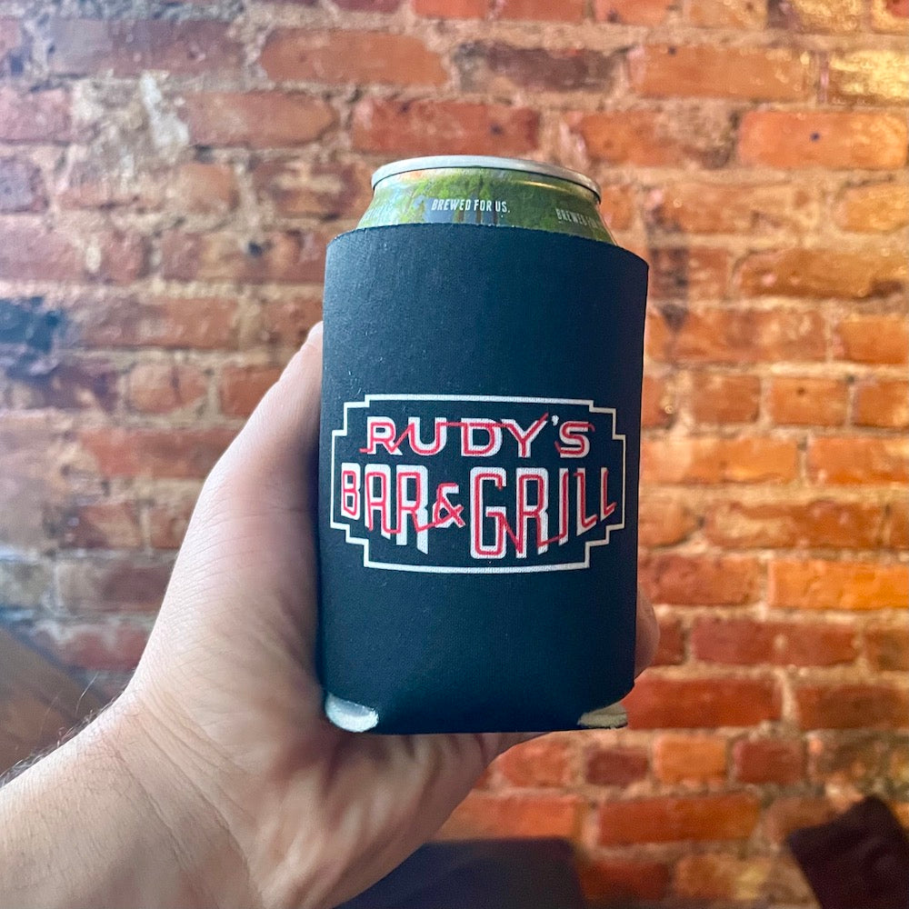 Hand holding Rudy’s Bar & Grill neon sign collapsible koozie with beer can in front of exposed brick wall at NYC bar