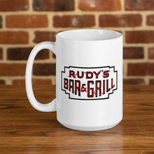 Load image into Gallery viewer, Close-up of a 15oz white mug featuring the Rudy’s Bar &amp; Grill neon sign logo, displayed on a wooden table with a blurred brick wall background.